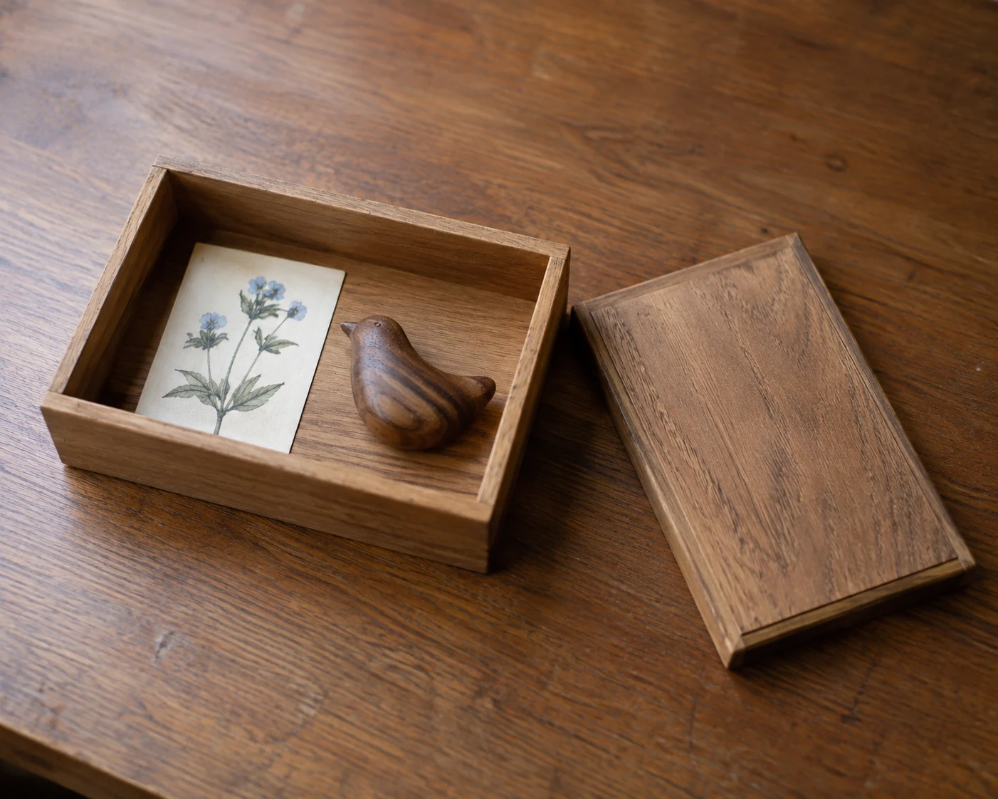 An open wooden box holds a botanical picture card and a wooden keepsake; its lid rests beside it.