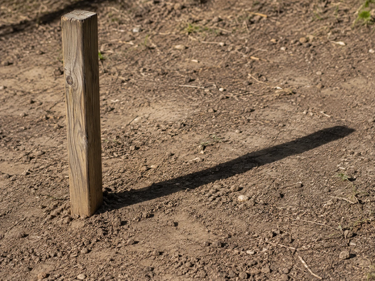 A wooden post casts a shadow across bare ground.