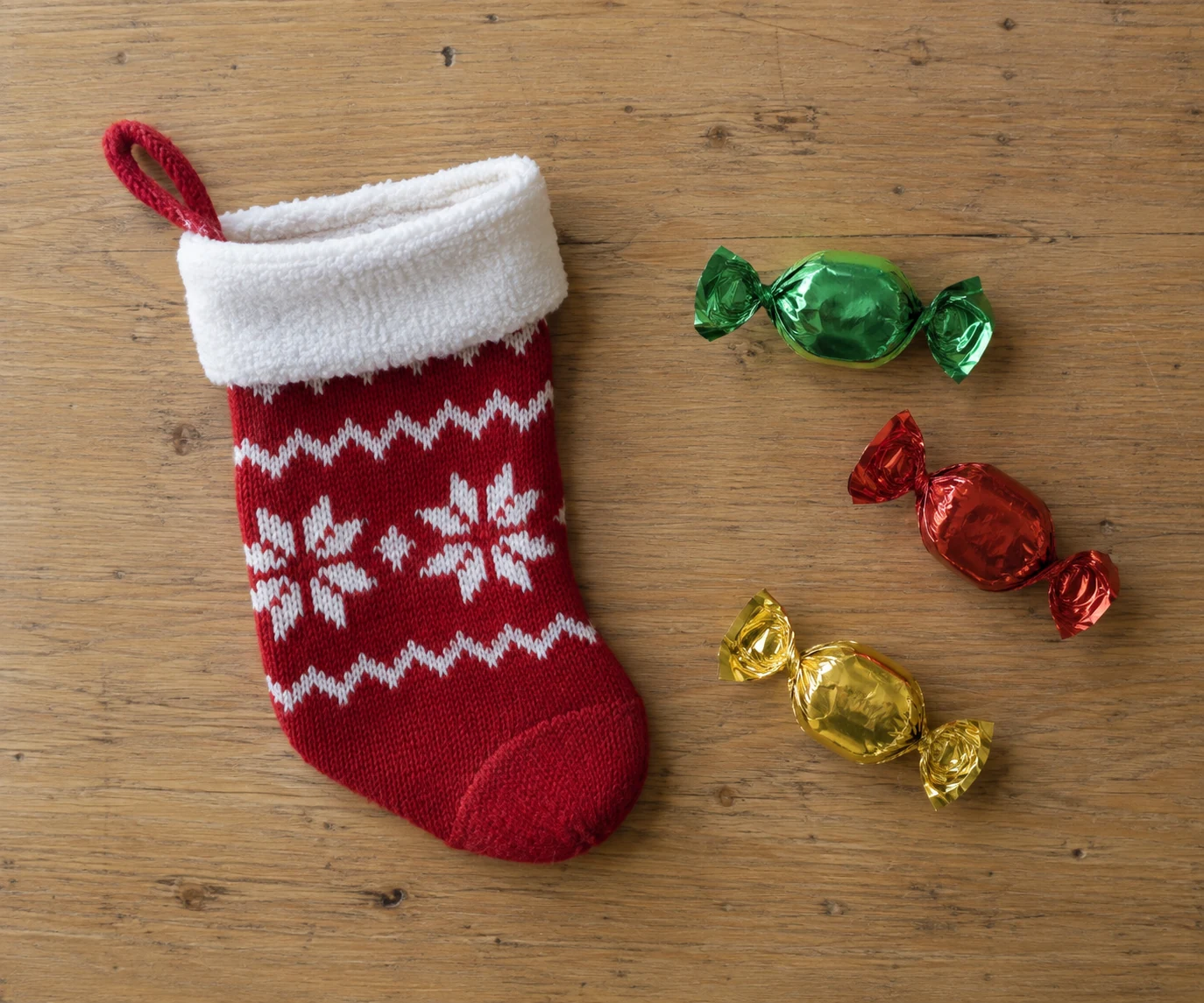 A small red knitted stocking lies on a wooden table beside three wrapped sweets.