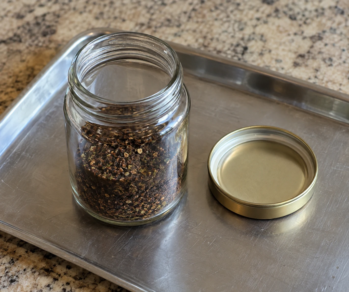 Open jar holding a dark dry blend, with its lid beside it on a protective tray.