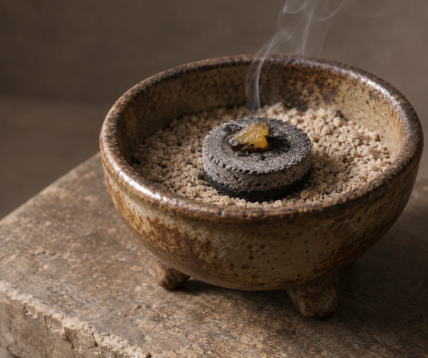 A small piece of resin sits on a grey charcoal tablet in a sand-filled ceramic burner resting on stone.