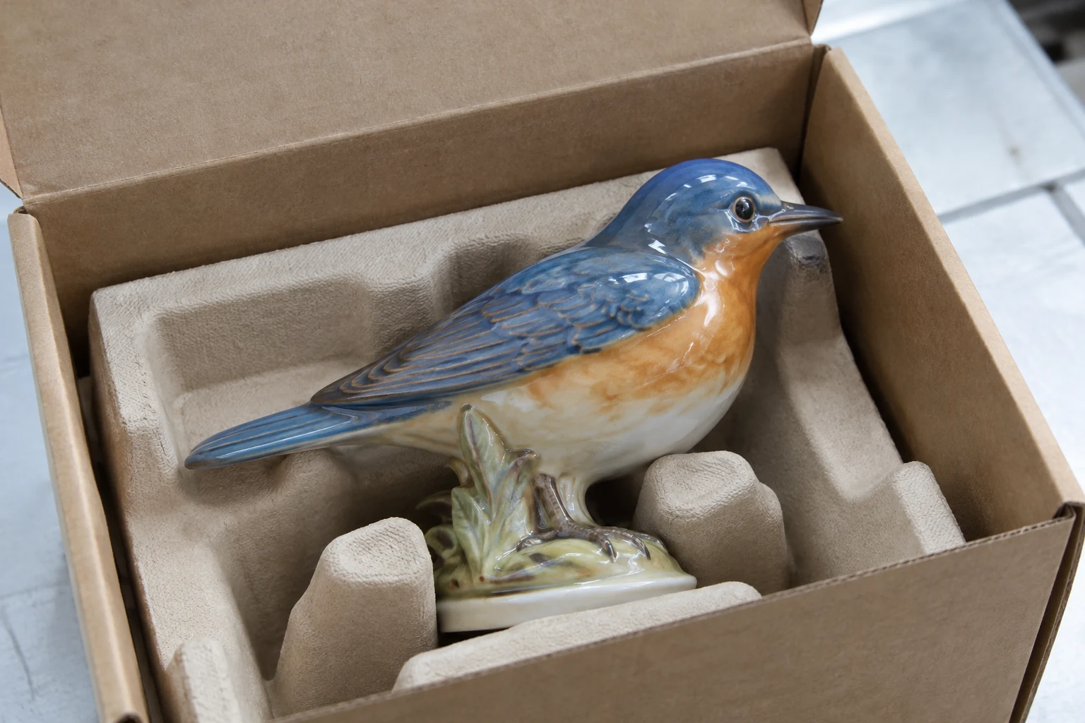 Ceramic bird figurine supported beneath its base in an open box, with space around its beak.
